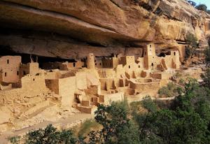 A cluster of tall buildings beneath an overhanging sandstone cliff. The buildings appear to be made of the same material as the cliff, and almost look like part of the landscape.