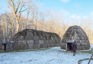 Two buildings made of wood, standing beside each other in a clearing. One of the buildings is short and round, while the other is long and narrow.