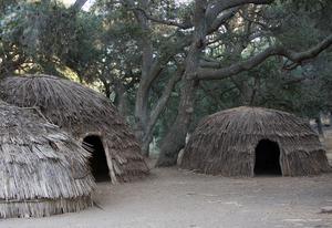 Three domed houses in a sandy clearing. Their entrances point towards each other, and their exterior walls appear to be covered in sticks.