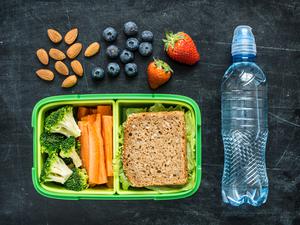 A photo of a school lunch. There is a bottle of water, almonds, blueberries, strawberries, and a container holding broccoli, carrots, and a sandwich.