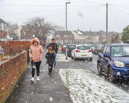 Snow falls from a gray sky. Three people in winter coats walk on the sidewalk.